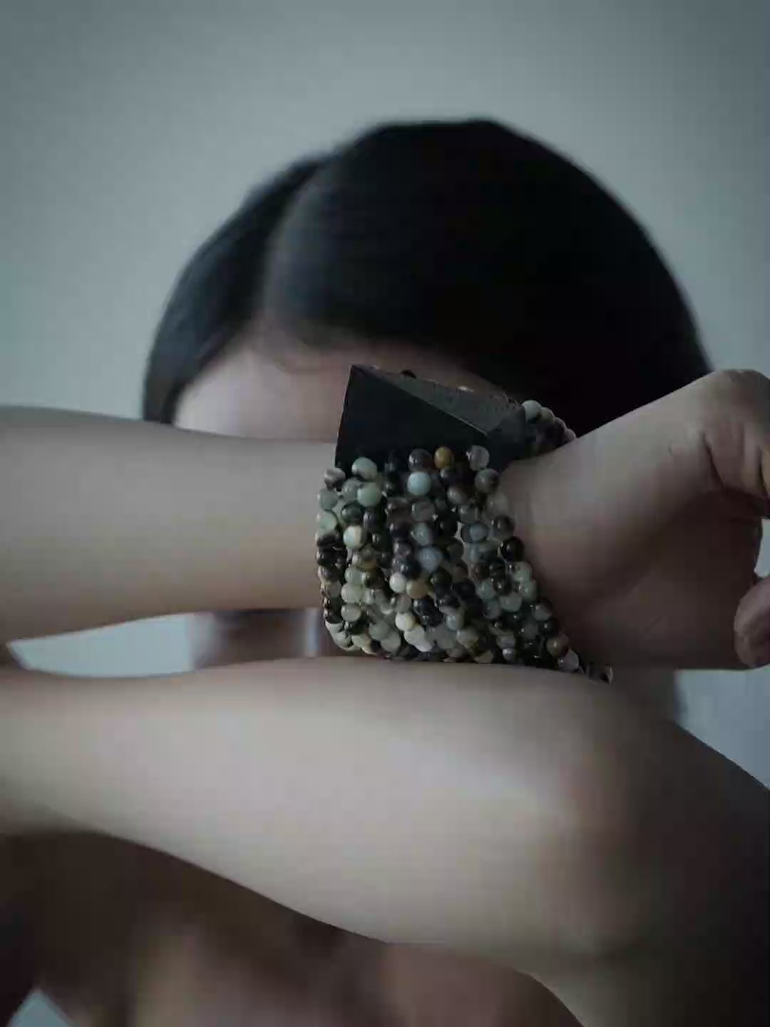 A close-up lifestyle shot of a model wearing a wide, multi-strand beaded cuff bracelet on her wrist, with her arms crossed over her face against a plain gray background.