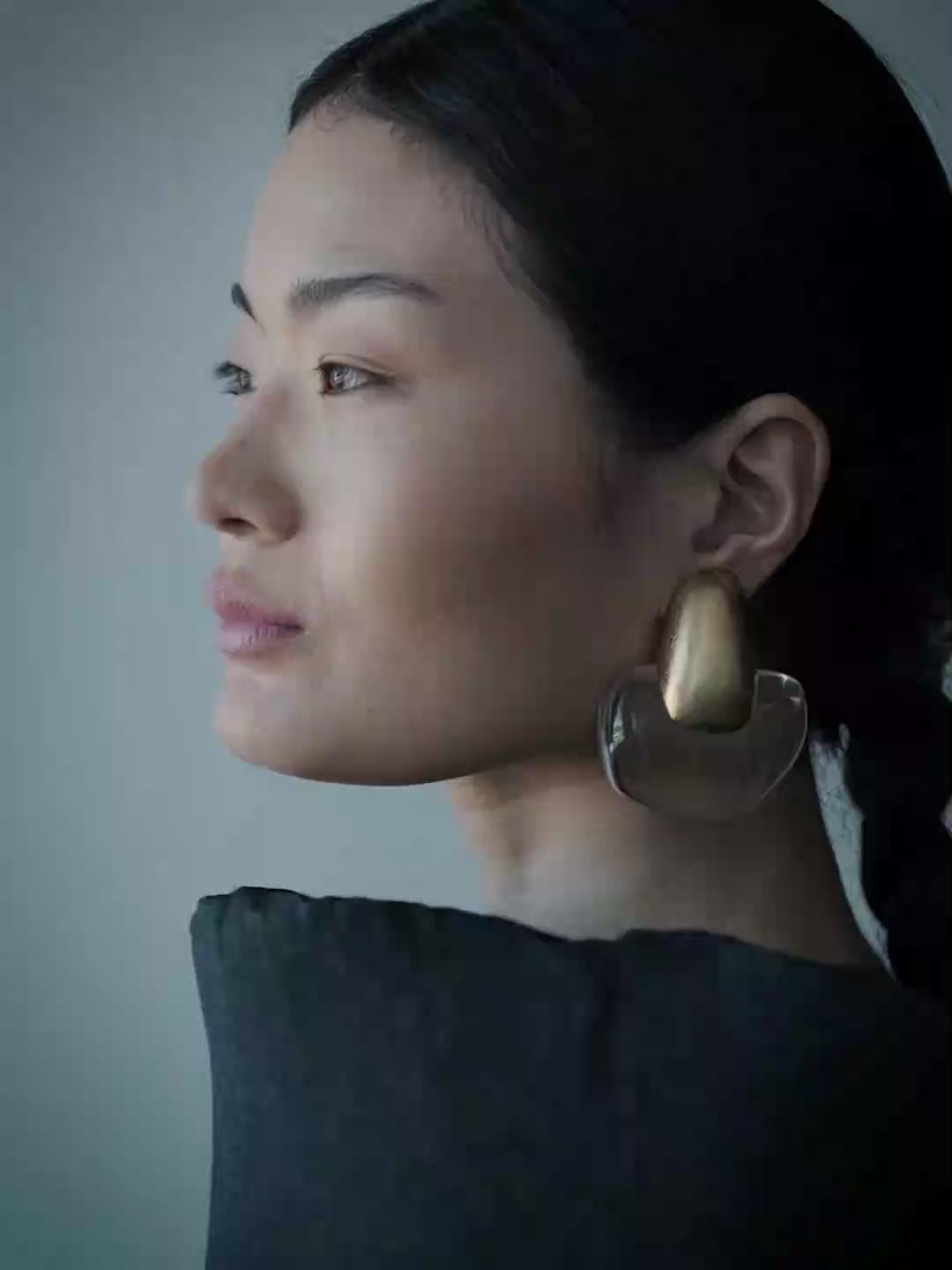 A close-up side profile of a woman wearing a large, sculptural statement earring. The earring features a textured, metallic gold top section and a clear, organic-shaped lower section.