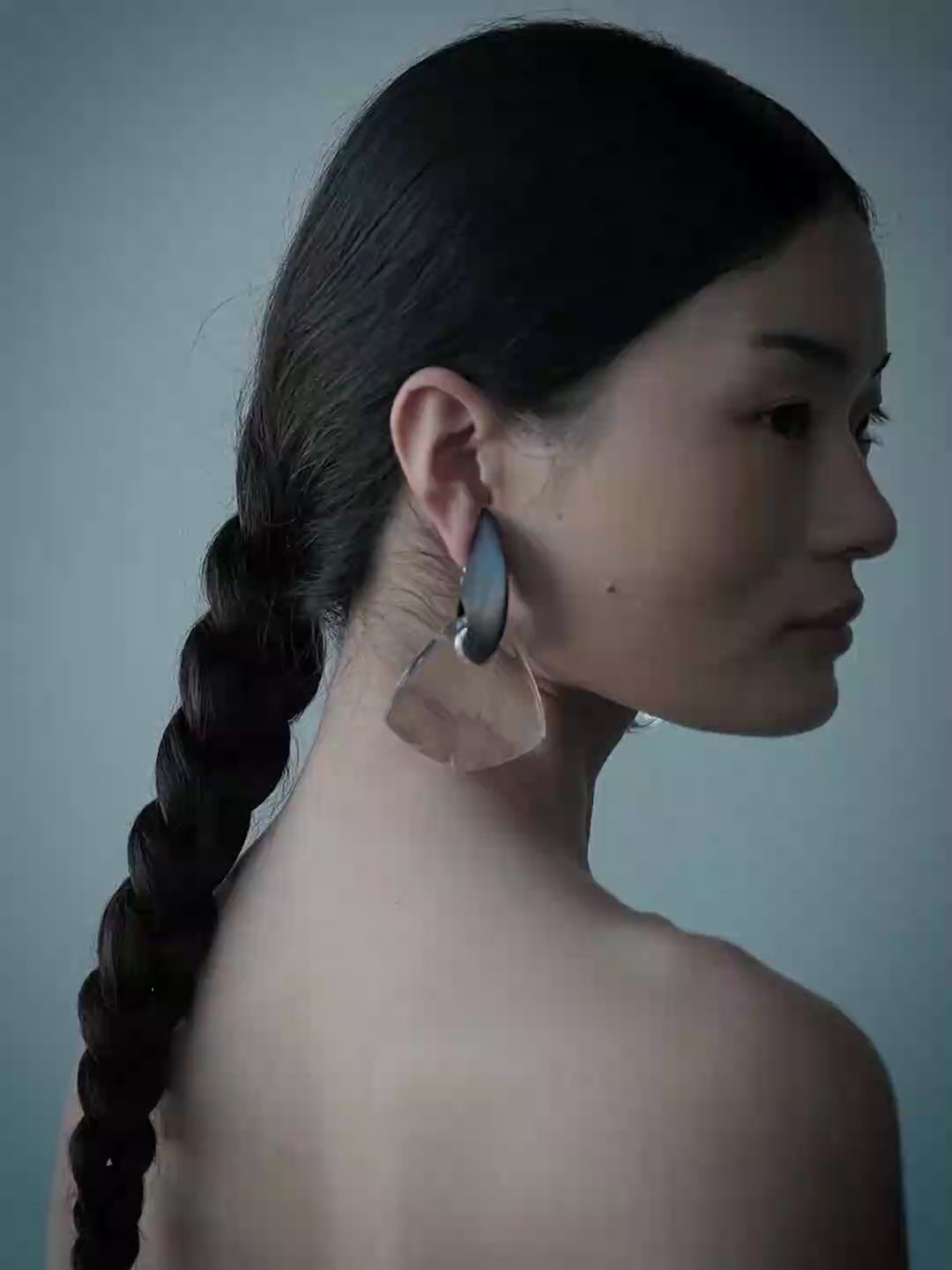 A woman wearing large, asymmetrical statement earrings made of layered translucent beige and dark gray materials, set against a solid blue-gray background.