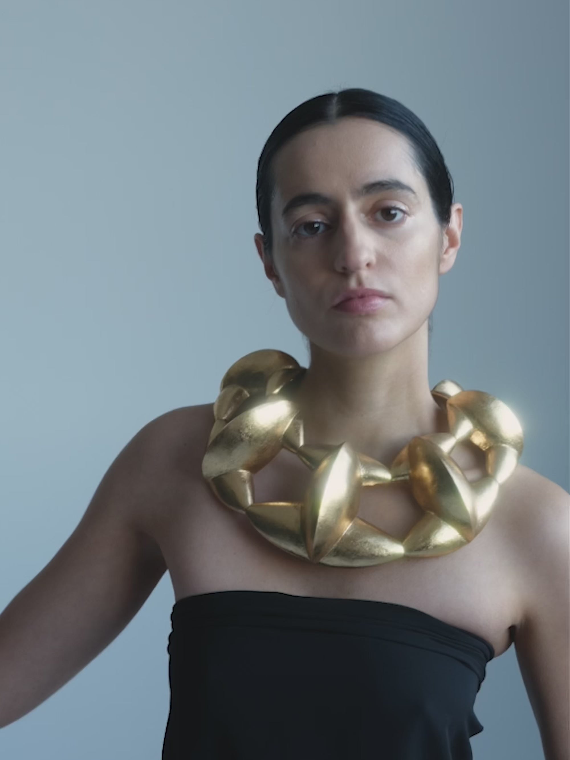 Woman wearing a black strapless dress and large gold necklace against a gray background