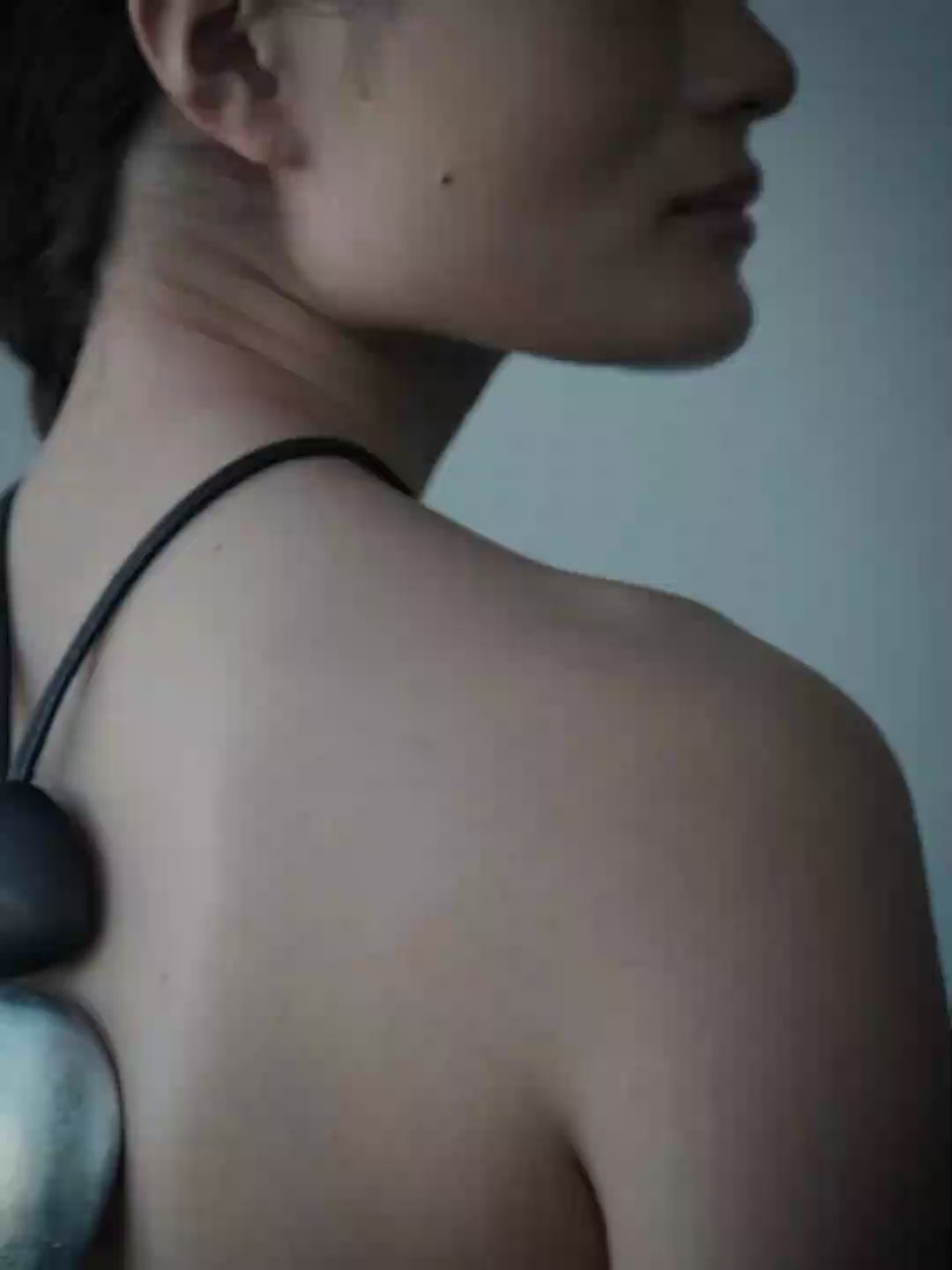 A woman shown from behind wearing a unique black cord pendant with two smooth pieces, one black and one silver, resting on her upper back.