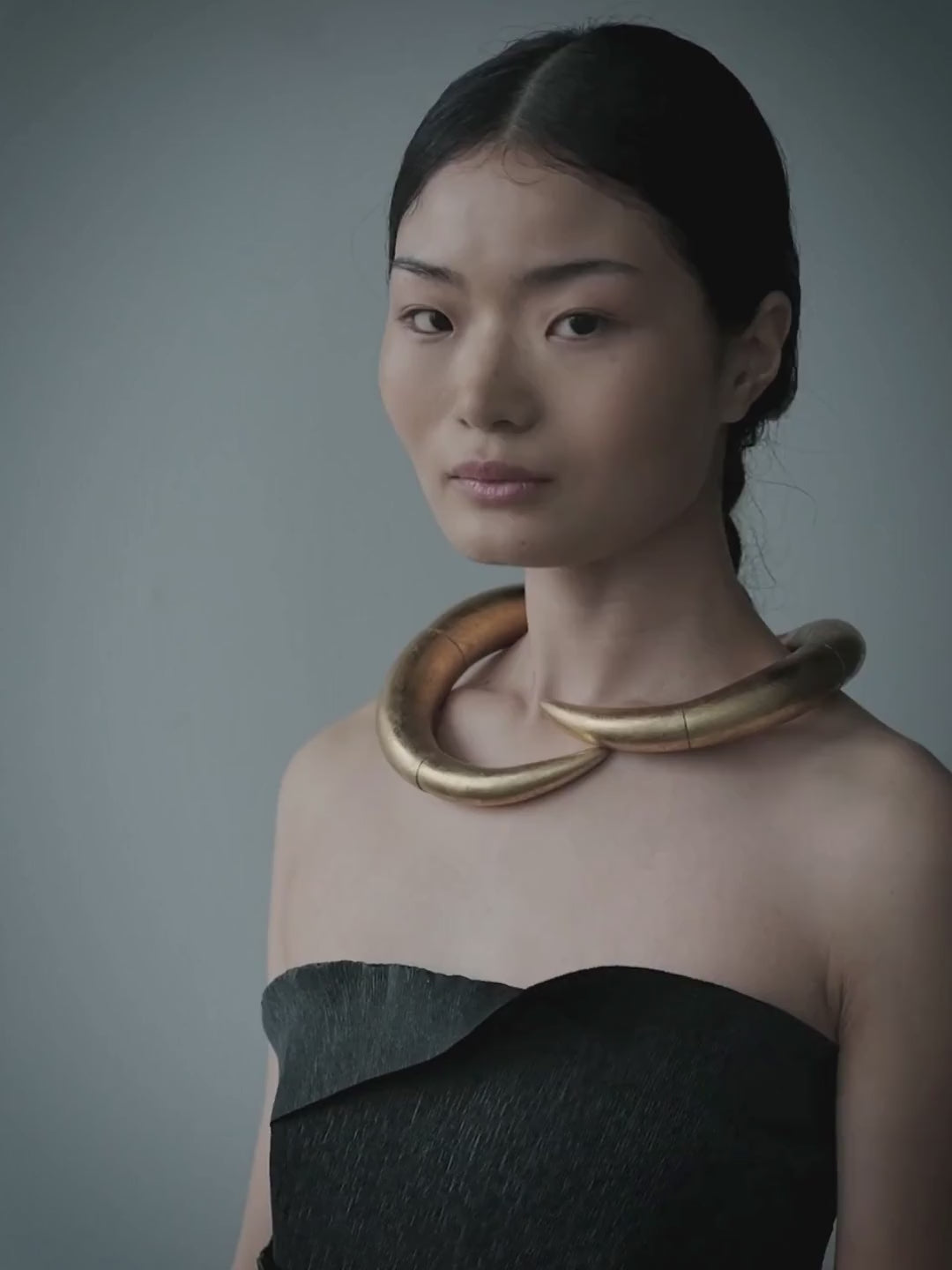 A model with dark hair pulled back wears a chunky, multi-strand gold collar necklace and a dark strapless top, posing with her arms outstretched against a solid slate blue-gray background.