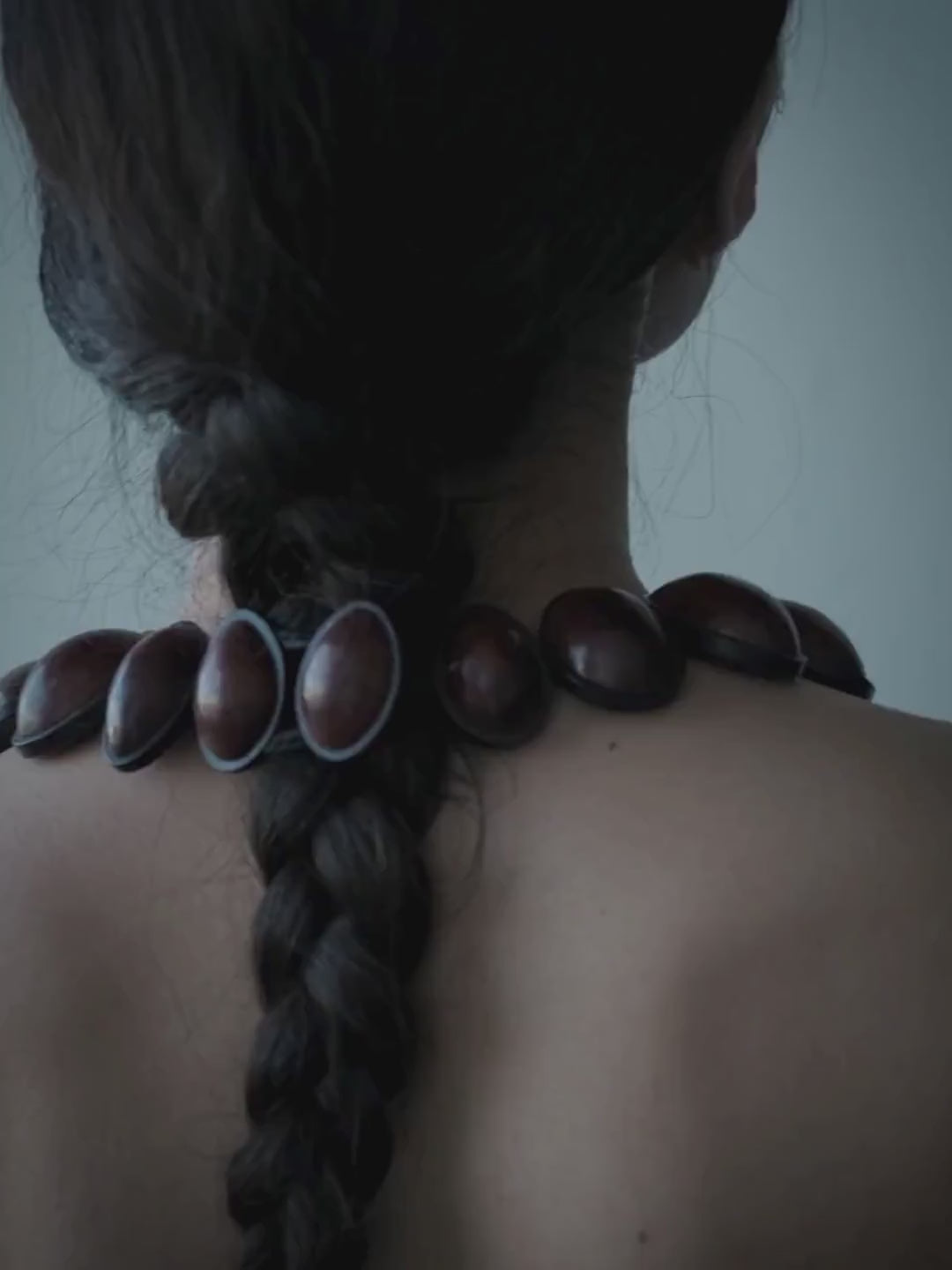 A close-up portrait of a woman wearing a chunky, dark reddish-brown wooden bead necklace against a muted blue-gray background.