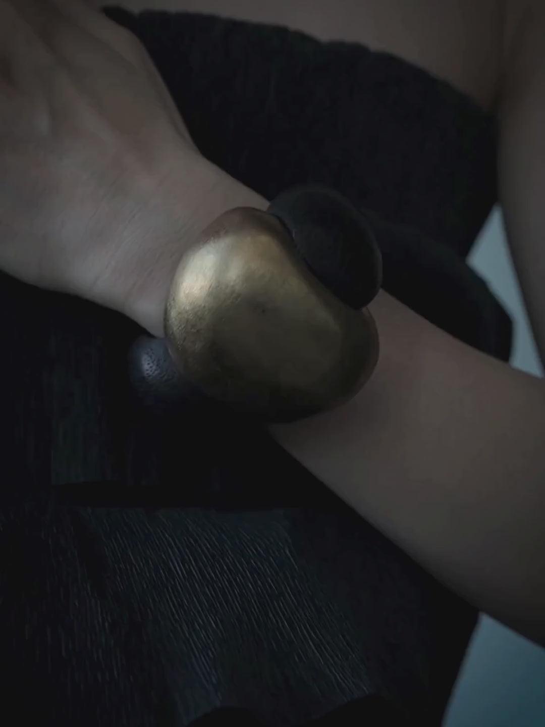 A woman wearing a chunky, sculptural gold and black cuff bracelet, posing with her hands on her neck against a solid blue-gray studio background.