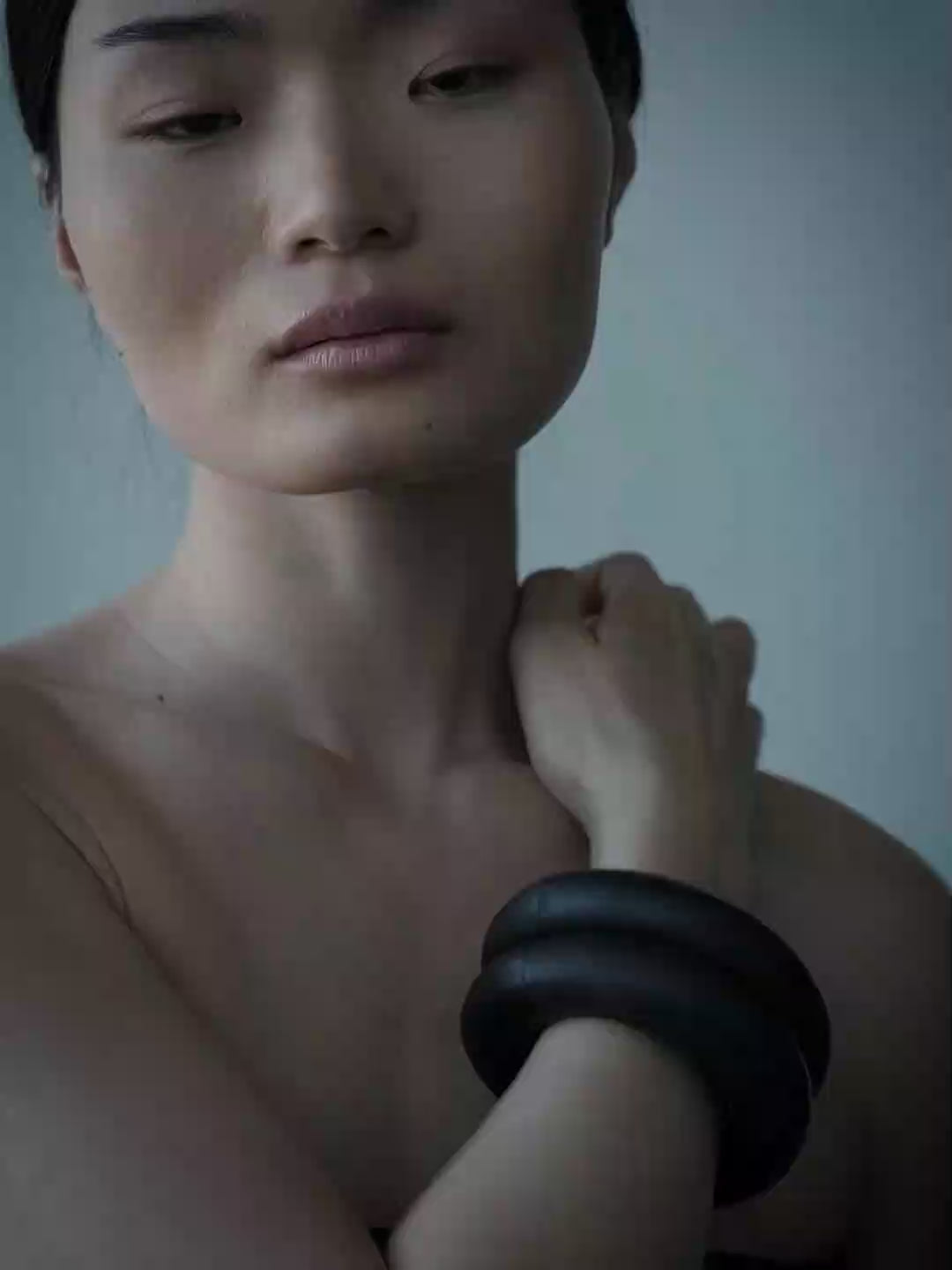 A woman with dark hair in a braid wearing a thick black circular wrist cuff, posed against a solid blue-gray background.