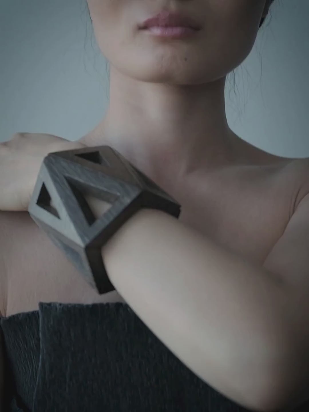 A woman with dark hair tied back wears a geometric wooden cuff bracelet on her raised arm, posed against a plain blue-gray background.