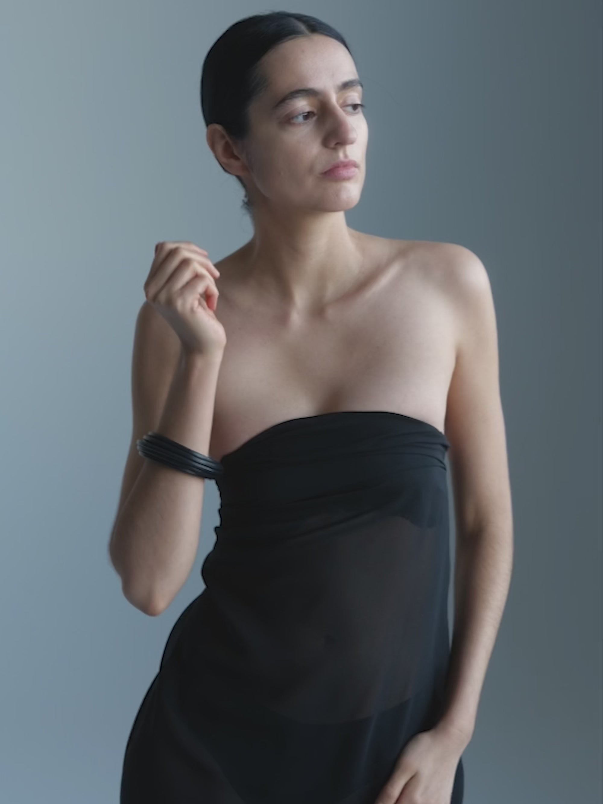 Woman in a black dress  wearing a black bracelet, against a gray background
