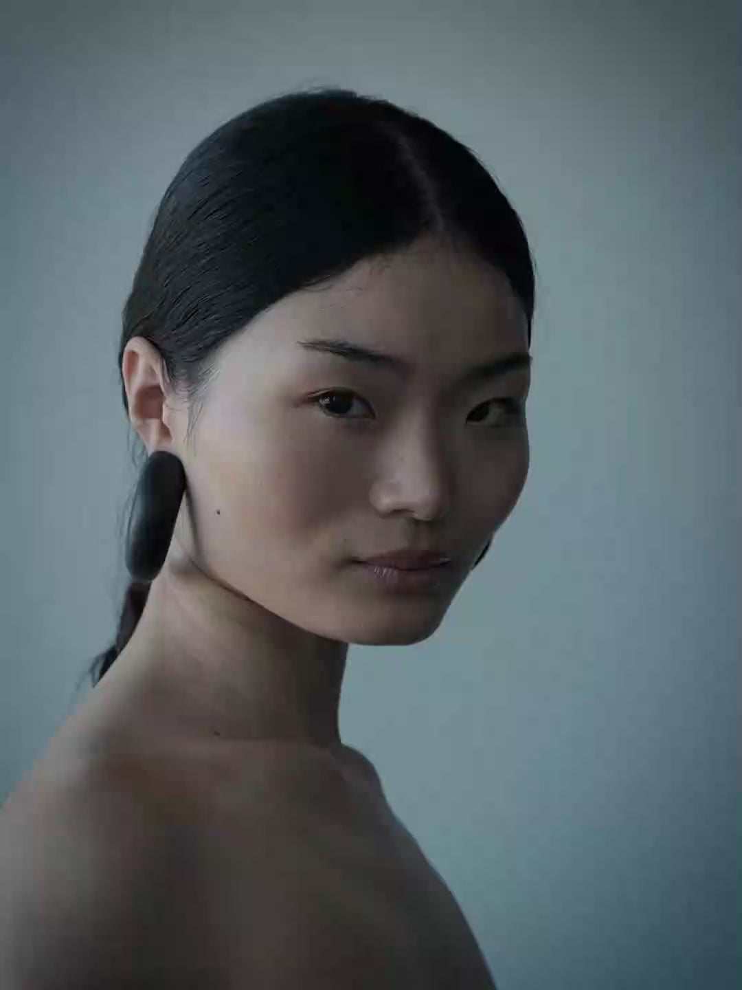 A woman in profile wearing a black strapless top and large dark wooden hoop earrings, photographed against a cool blue-gray studio background.