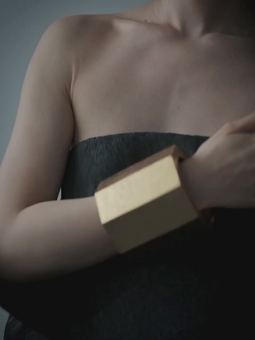 A woman with her hair pulled back wears a wide, chunky gold and black cuff bracelet on her raised arm, posing against a muted slate blue background.
