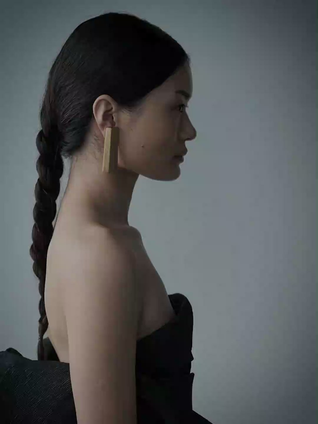 A woman in profile wearing a black strapless dress and a gold rectangular earring, standing against a plain light gray wall.