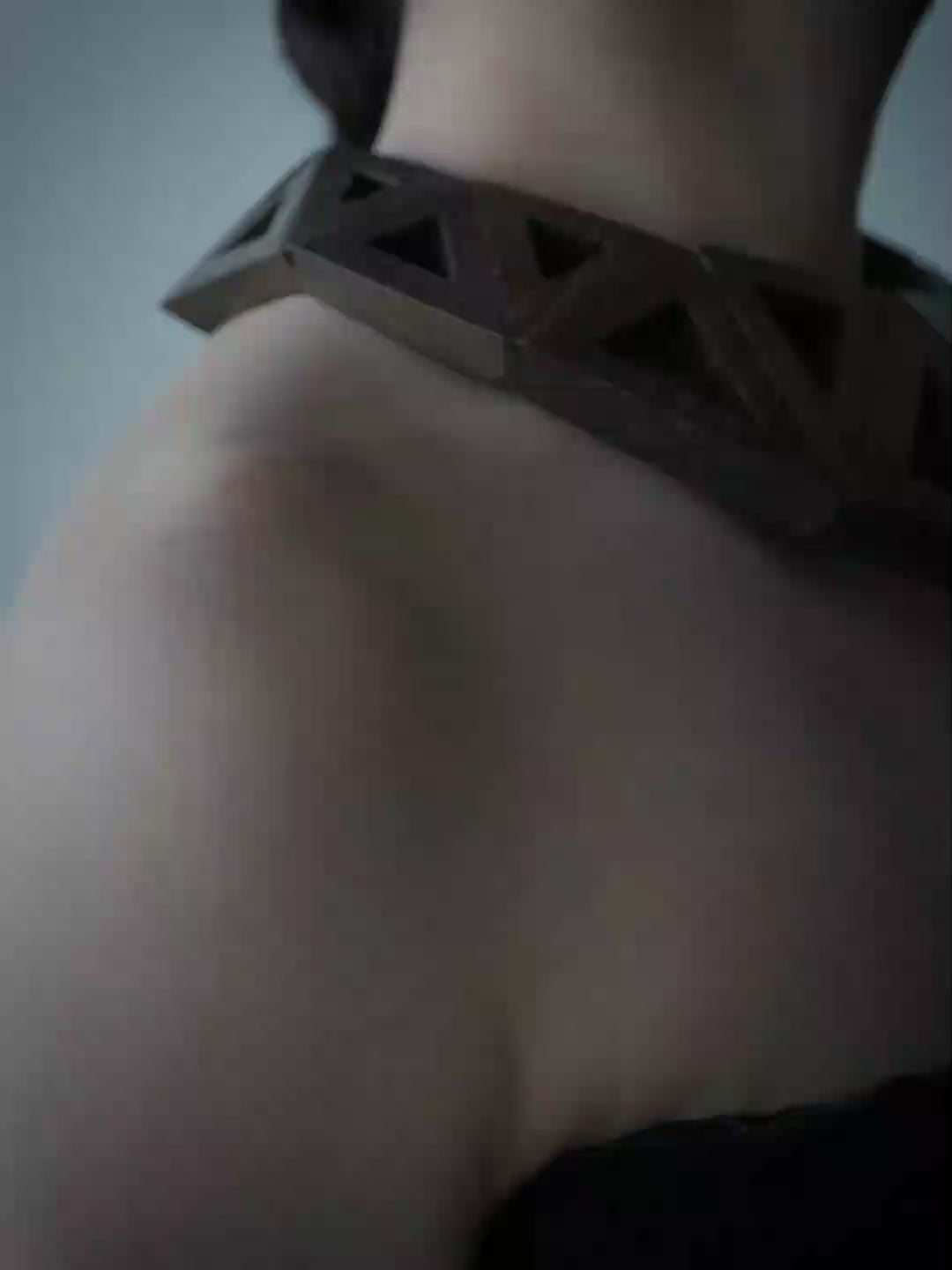A woman with dark hair pulled back wears a large geometric dark wood collar necklace against a plain blue-gray background.
