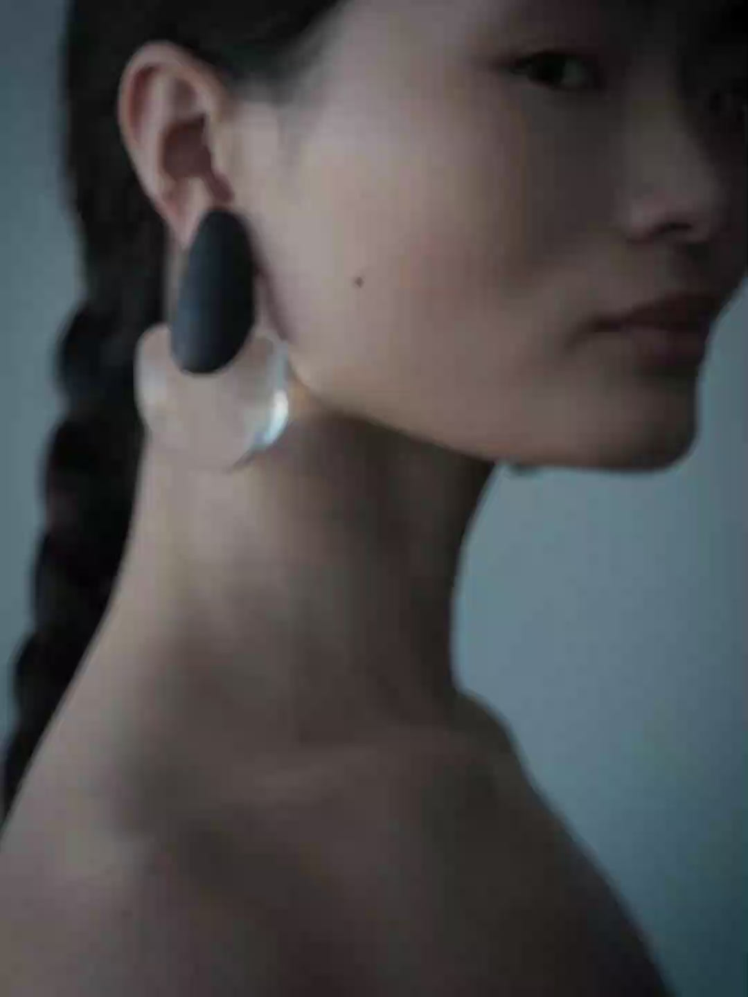 A close-up side profile of a woman wearing a large, sculptural statement earring. The earring features a dark navy blue top section and a large, organic, translucent pale pink lower section.