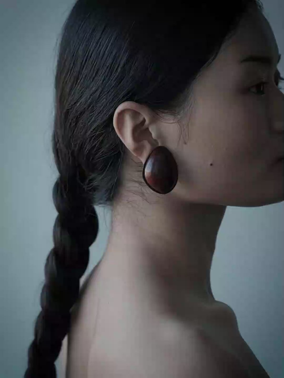 A woman in a black strapless dress wearing large dark circular earrings, photographed against a muted blue-gray background.
