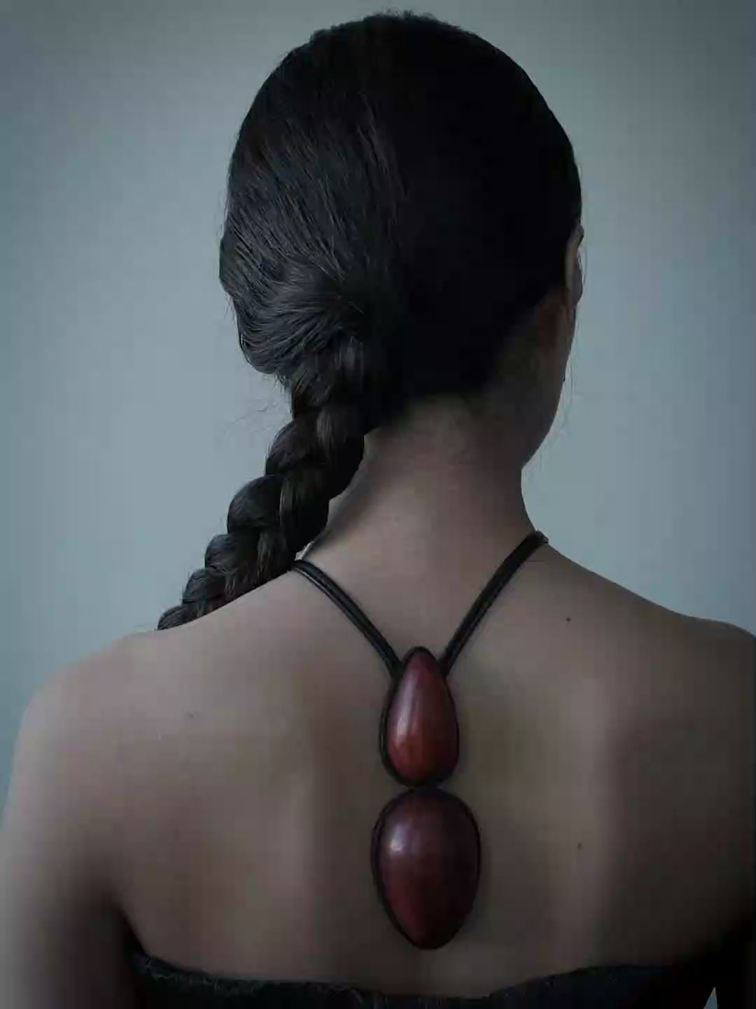 A woman viewed from behind wearing a black strap pendant with two large, polished dark red wooden beads, set against a plain blue-gray wall.