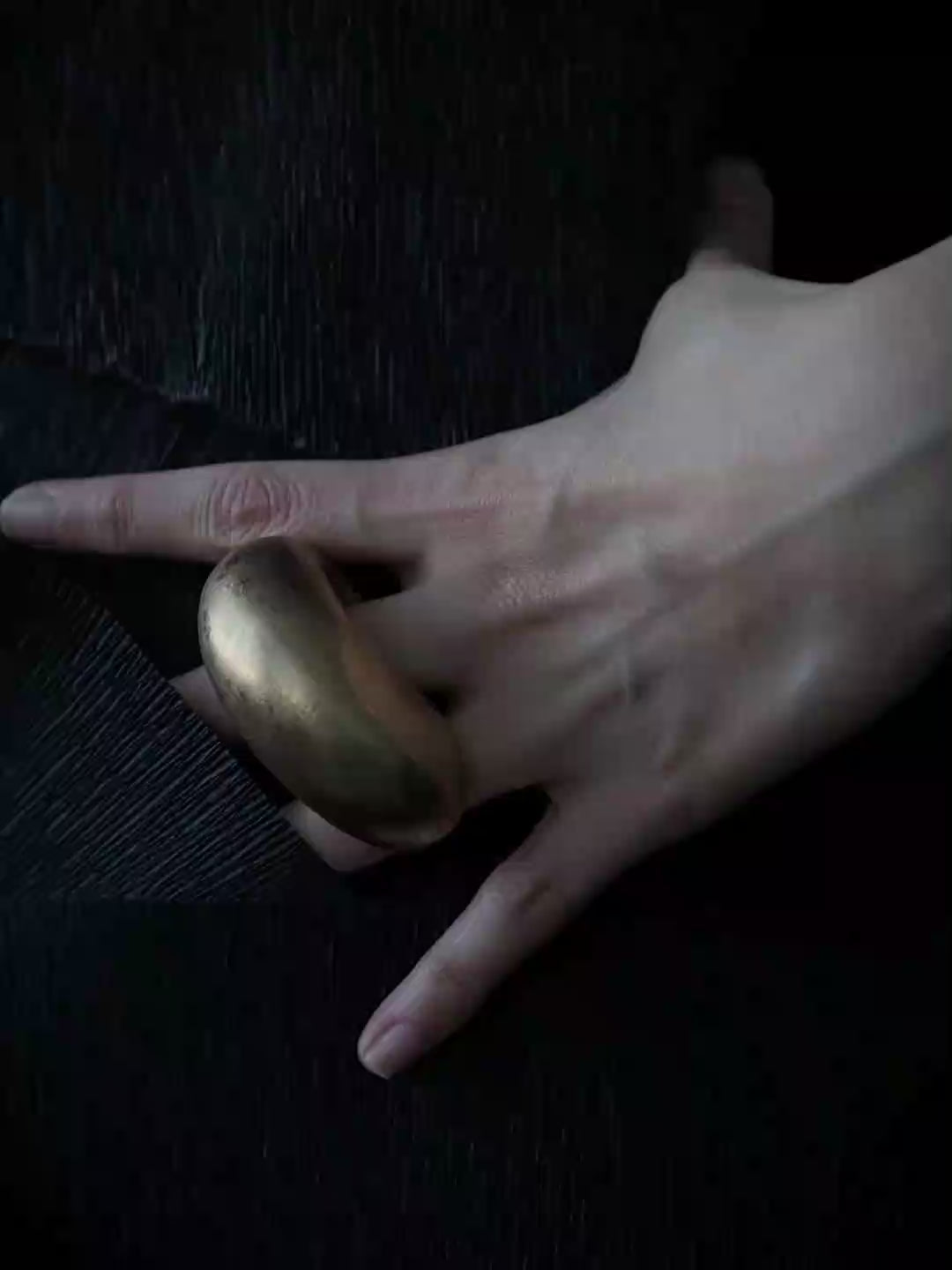A model with dark hair pulled back wears a large, smooth, matte gold oval ring on her finger, posing against a solid slate blue-gray background.