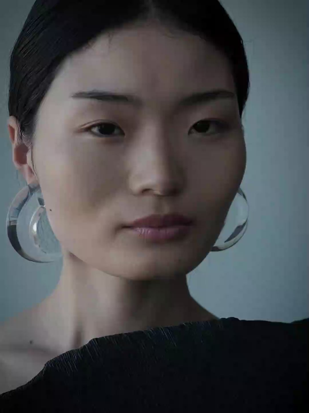 A close-up side profile of a woman with dark hair in a braid, wearing a large, chunky clear acrylic hoop earring against a muted blue-gray background.