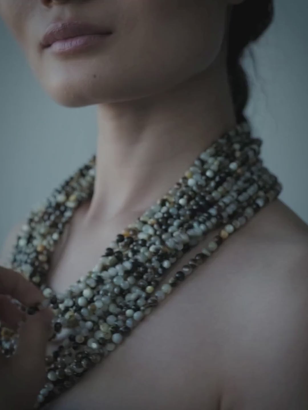 A model with dark hair pulled back wears a multi-strand beaded pearl necklace with metallic gold, silver, and green beads, paired with a dark strapless top against a cool blue-gray background.