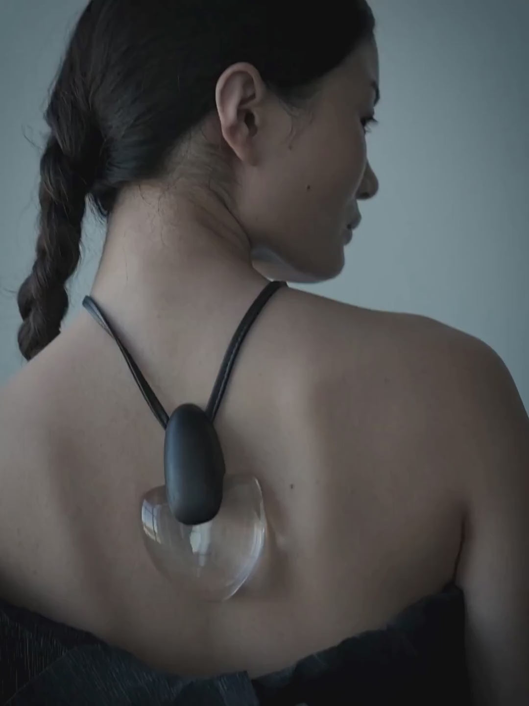 A woman shown from the back wearing a black strap necklace with a large, organic-shaped clear glass or acrylic pendant. She is wearing a black off-the-shoulder top against a cool, blue-gray studio background.