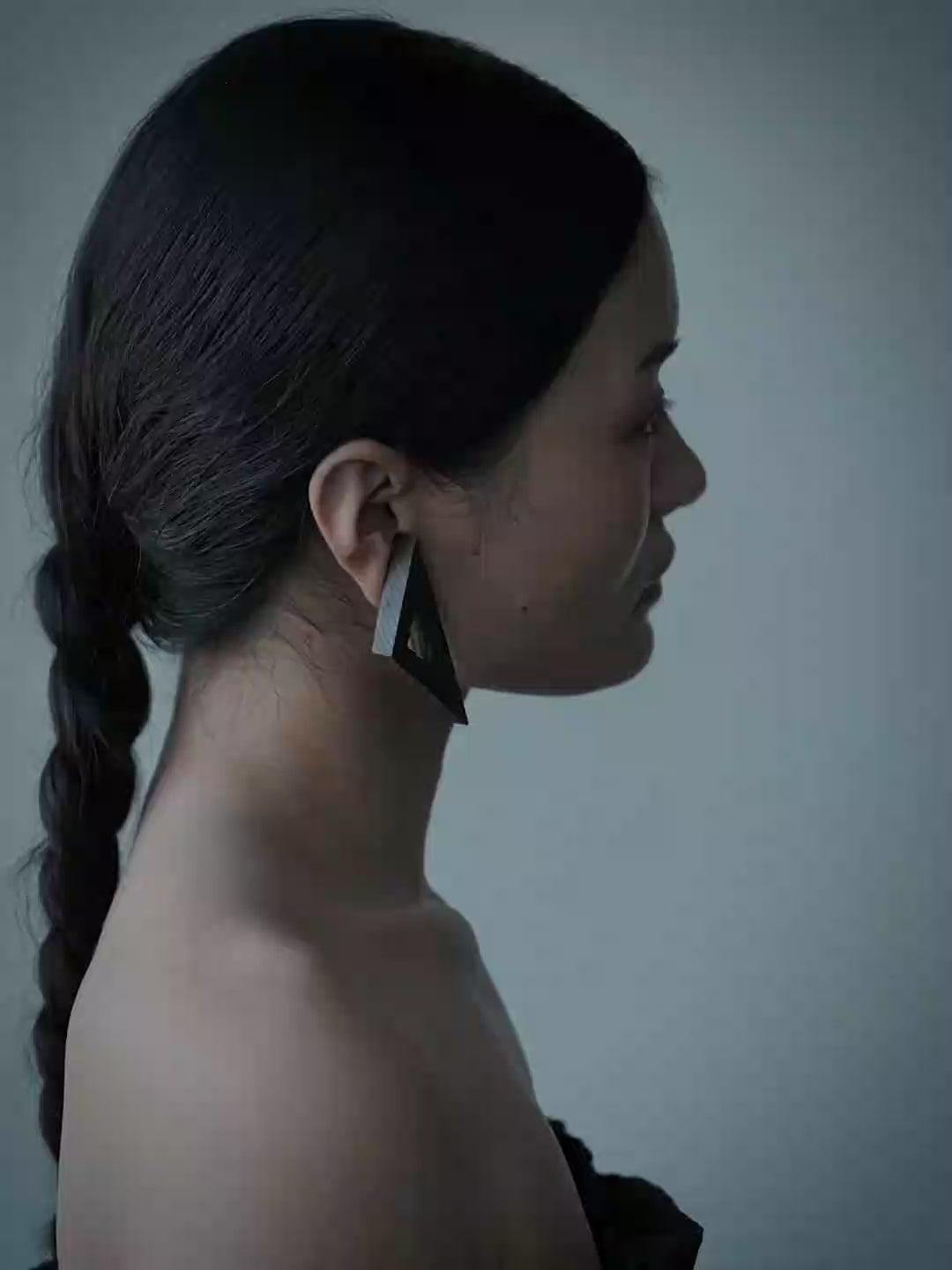 A close-up portrait of a woman with dark hair pulled back, wearing a large geometric wooden earring. The background is a solid, muted slate blue-gray.