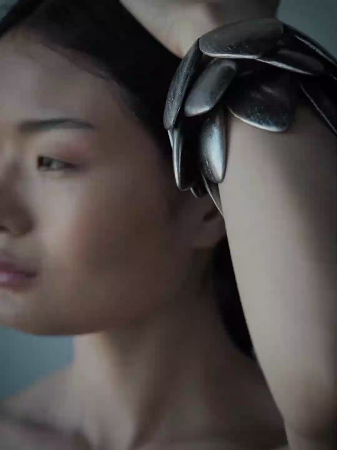 A close-up lifestyle shot of a person wearing a chunky, multi-layered metallic silver cuff bracelet on their wrist, with their hand resting on their head against a solid blue-gray background.