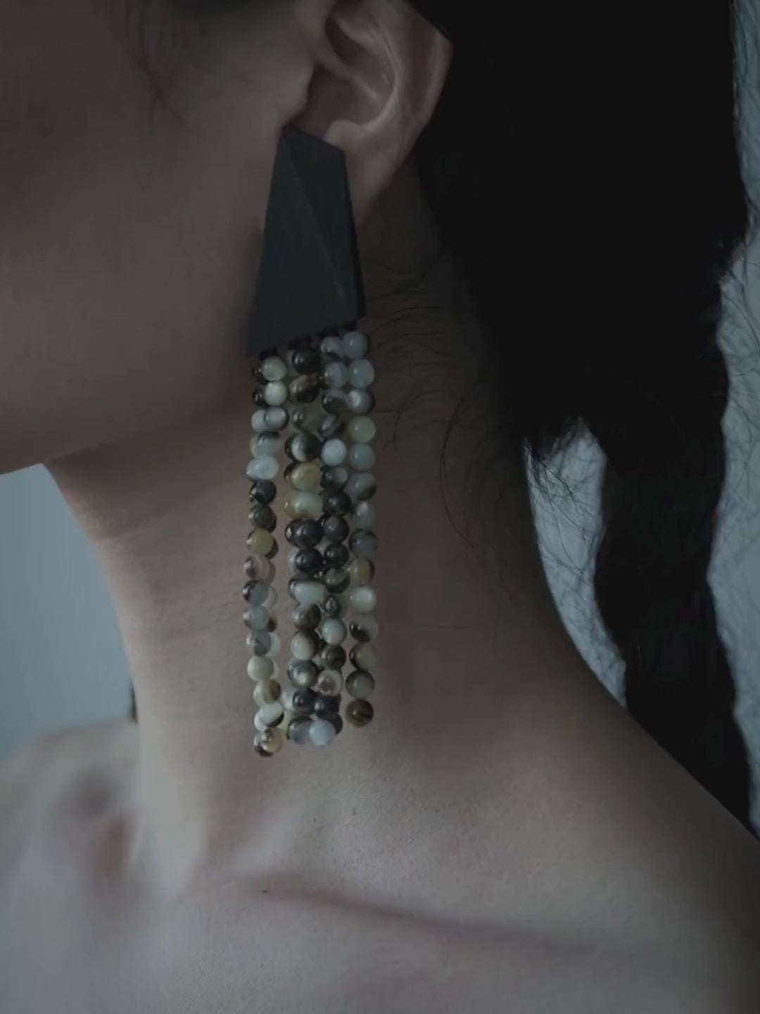 A woman in profile wearing long, statement drop earrings with green, gold, and dark elements, styled with a black strapless top against a muted blue-gray background.