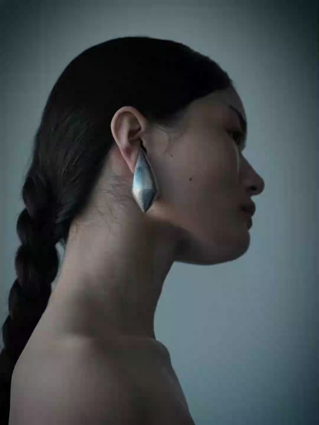 A side-profile portrait of a woman with dark hair in a braid, wearing a large, faceted silver geometric earring against a muted blue-gray background.