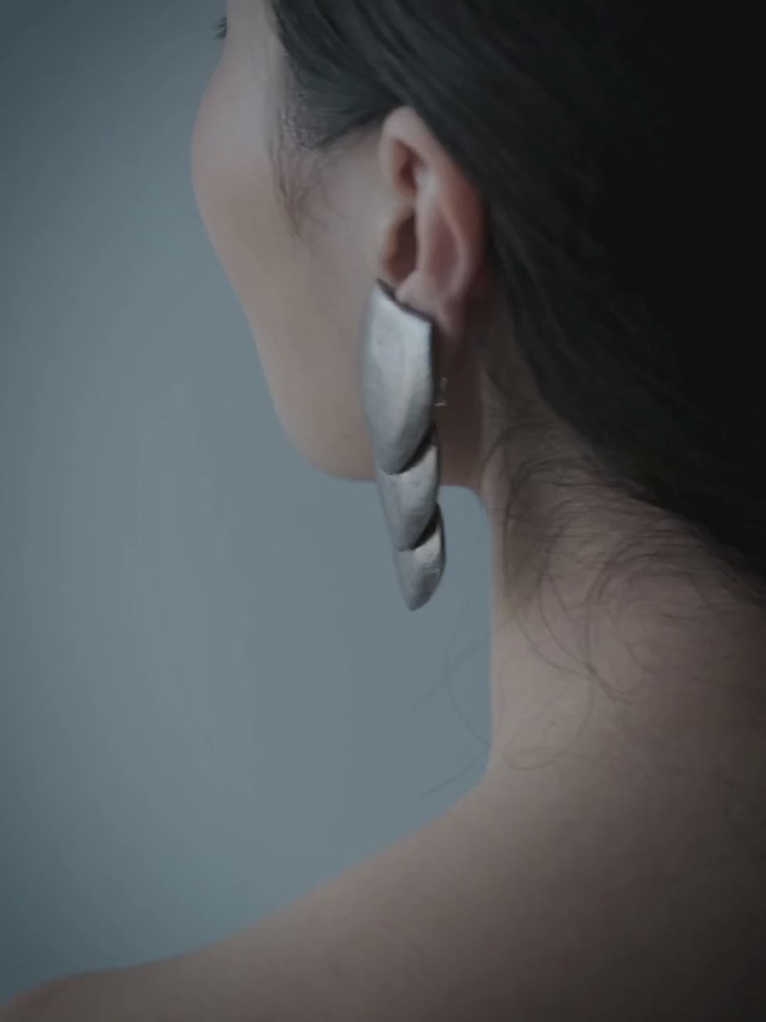 A woman with dark hair pulled back wears a large, sculptural white shell-shaped earring. She is wearing a dark strapless top against a plain, cool gray-blue background.