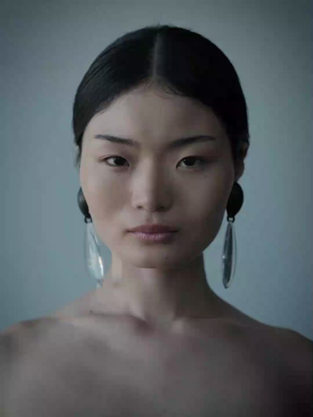 A woman wearing a large, asymmetrical teardrop earring made of clear glass or acrylic, paired with a black bralette, set against a solid blue-gray background.