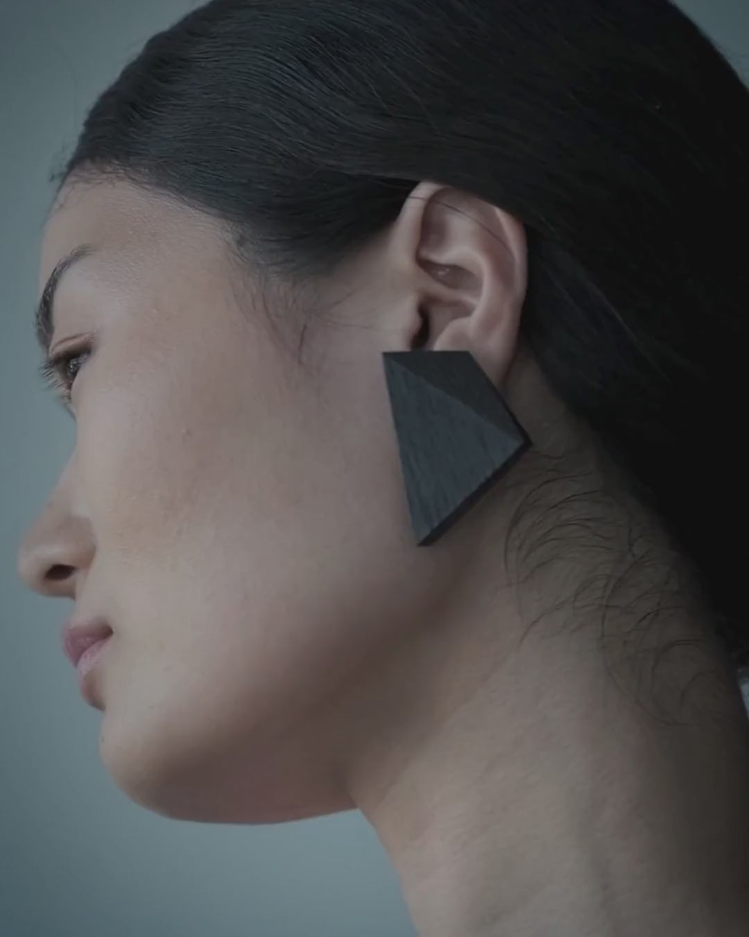 A close-up video of the side profile of a woman with dark hair in a braid, wearing a large, matte black geometric drop earring against a muted blue-gray background.