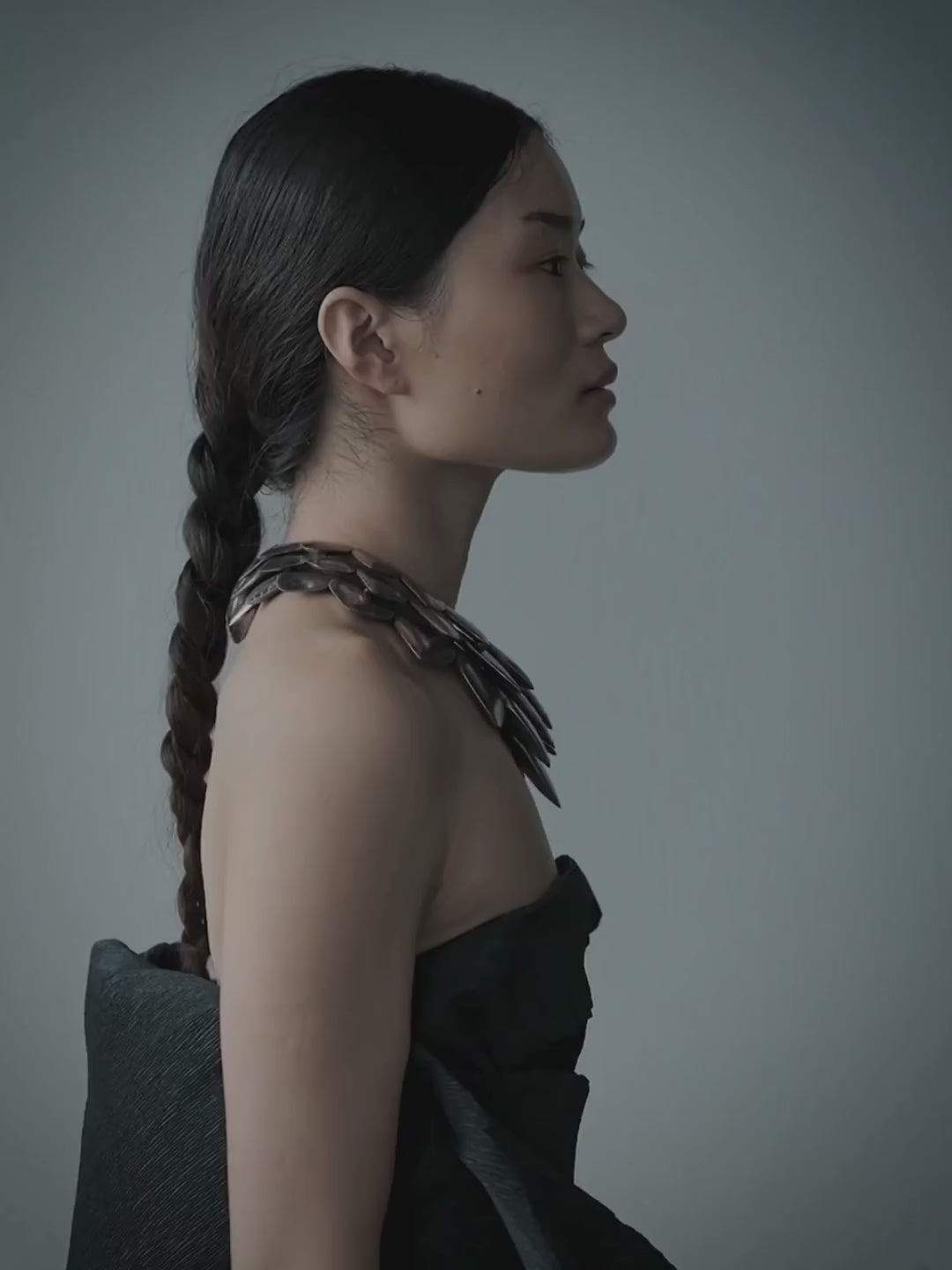 A model with dark hair tied back wears a dramatic black strapless dress and a bold geometric silver and black collar necklace, photographed against a plain gray studio background.