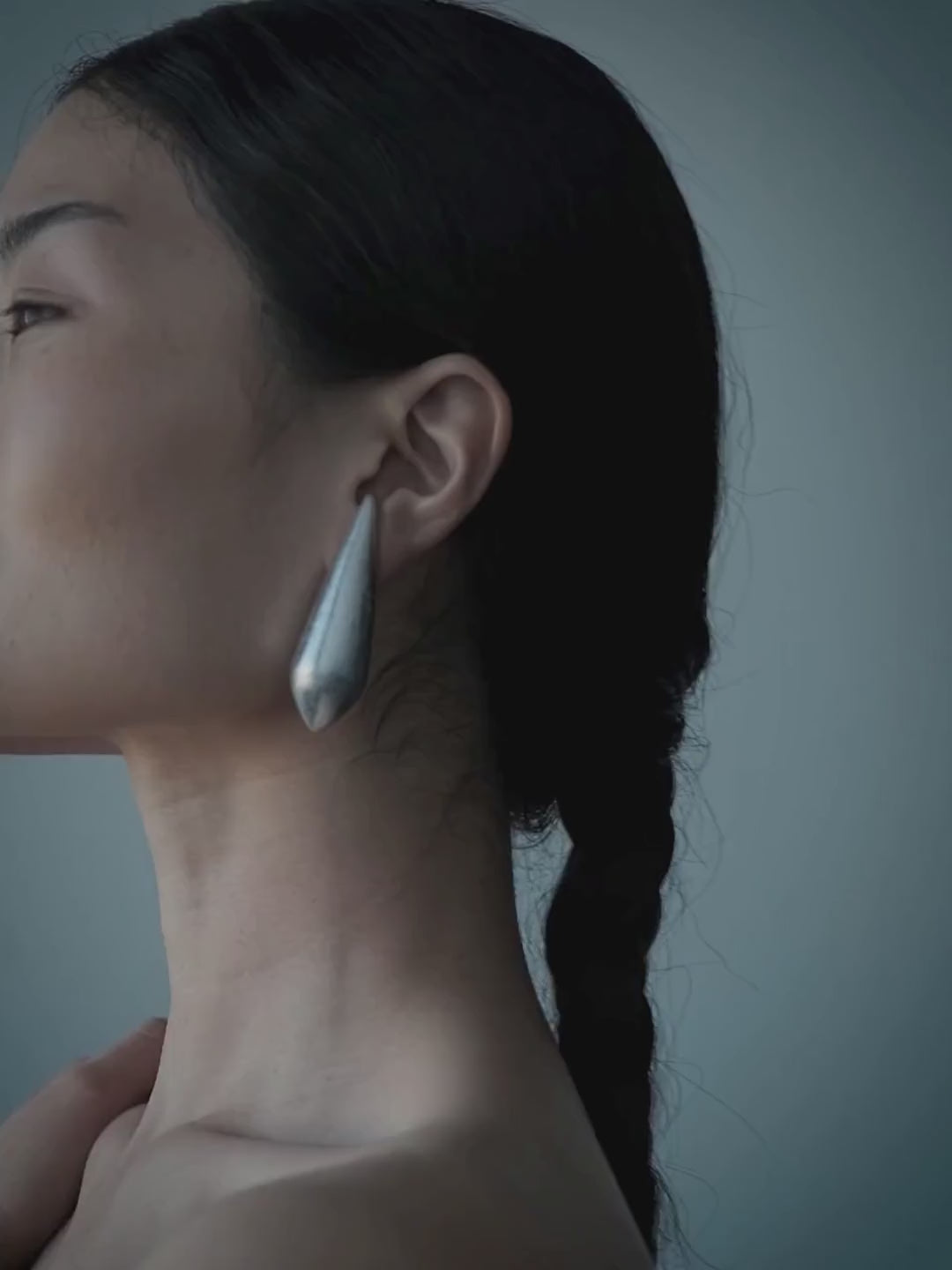 A woman in profile wearing a large, sculptural silver teardrop earring against a solid blue-gray background.