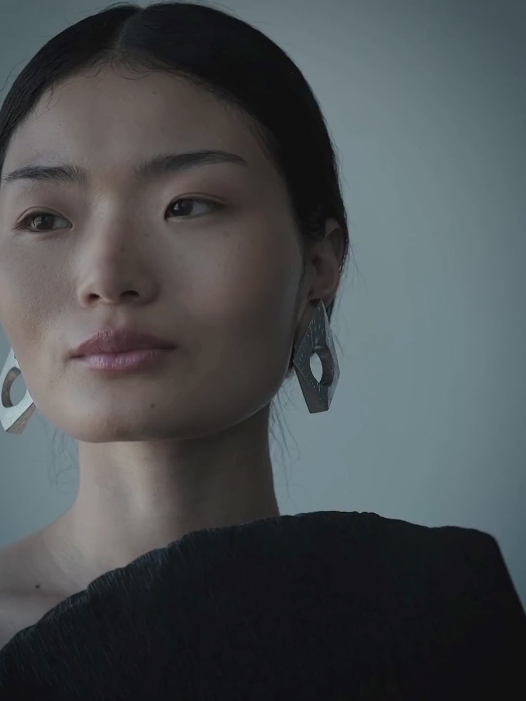 A woman wearing a large, geometric silver earring with a circular cutout, set against a dark textured top and a muted blue-gray background.