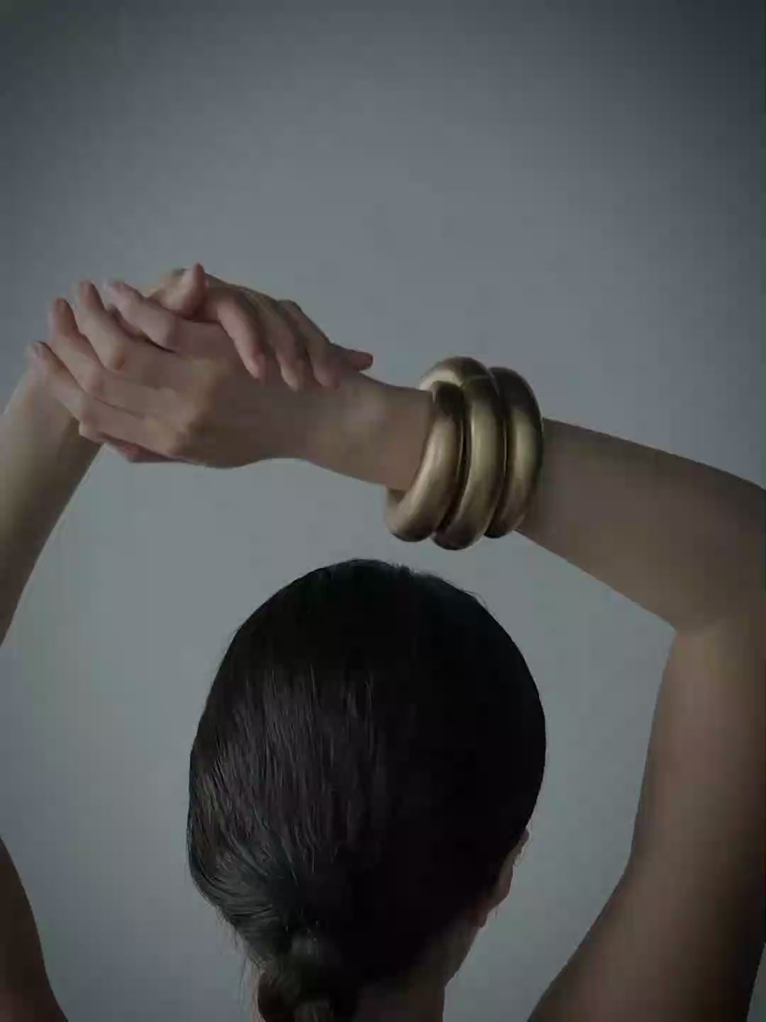 A woman viewed from behind with her arms raised, wearing a set of thick, stacked gold cuffs on her right wrist, against a solid blue-gray background.