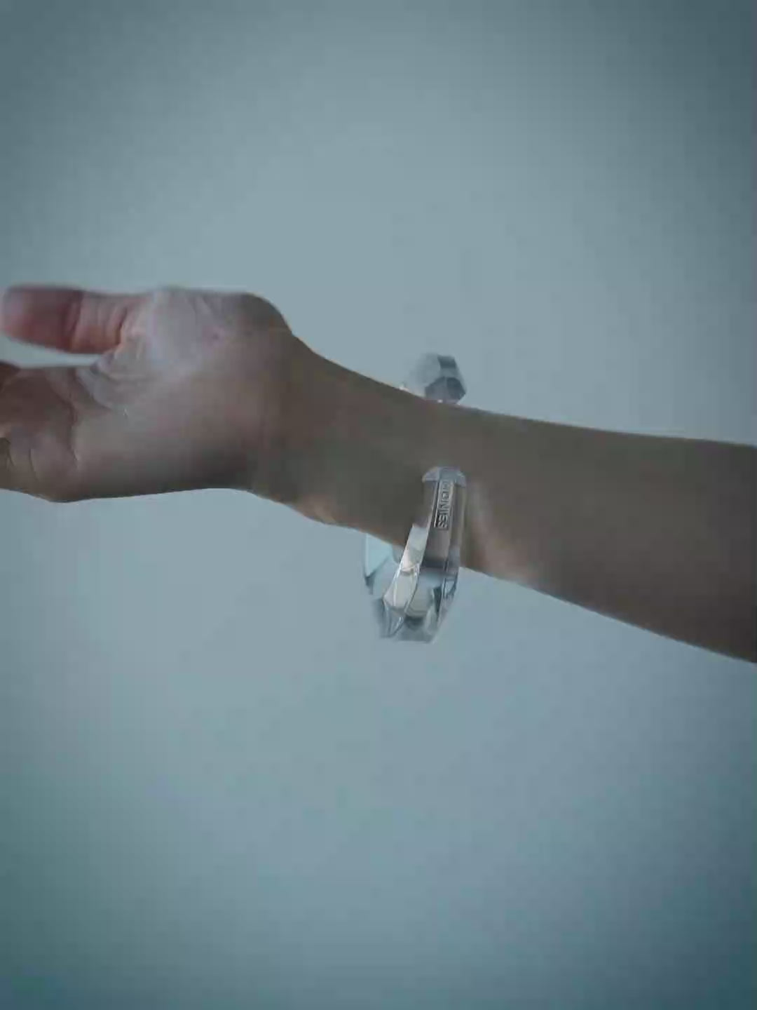 A model wearing a transparent bangle bracelet, shown from the chest up against a cool blue-gray background.