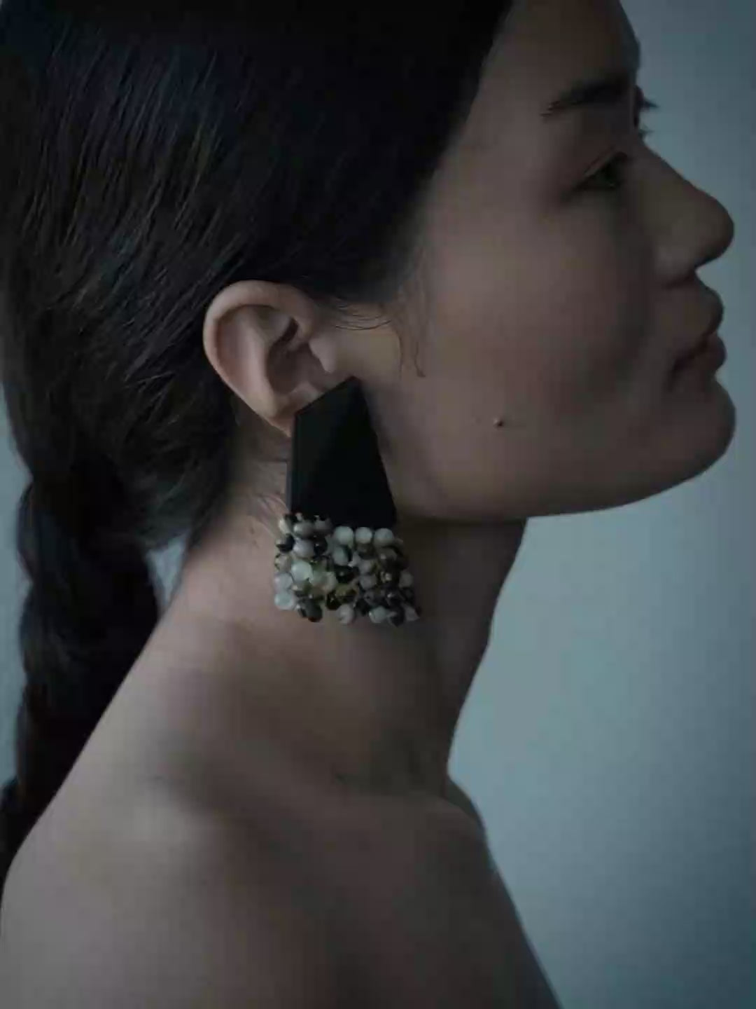 A close-up portrait of a model wearing a 
statement earring with a dark gray textured panel and cascading beads in black, white, and gold, set against a solid blue-gray background.