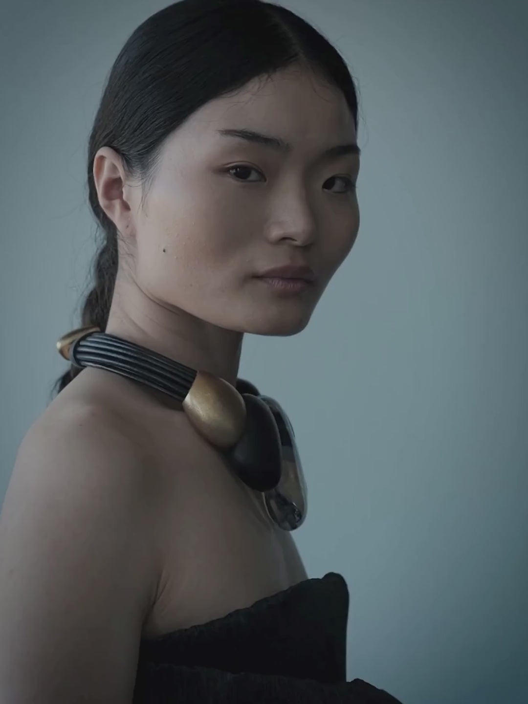 A woman with dark hair in a braid wears a chunky, sculptural collar necklace with gold, black, and translucent clear elements, paired with a black strapless top against a muted blue-gray background.