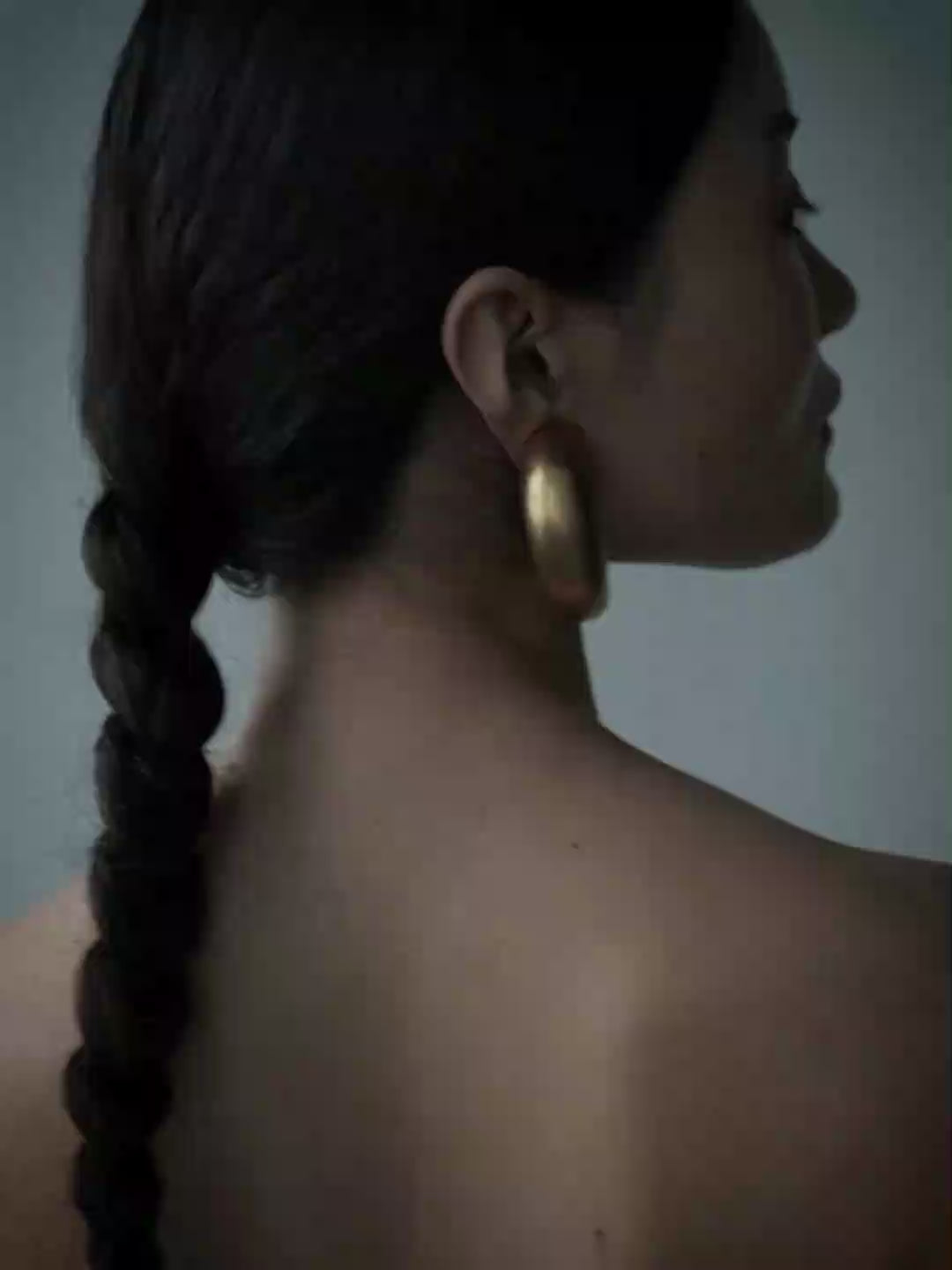 A woman with dark hair pulled back wears a strapless black dress and large gold hoop earrings, photographed against a plain blue-gray studio background.