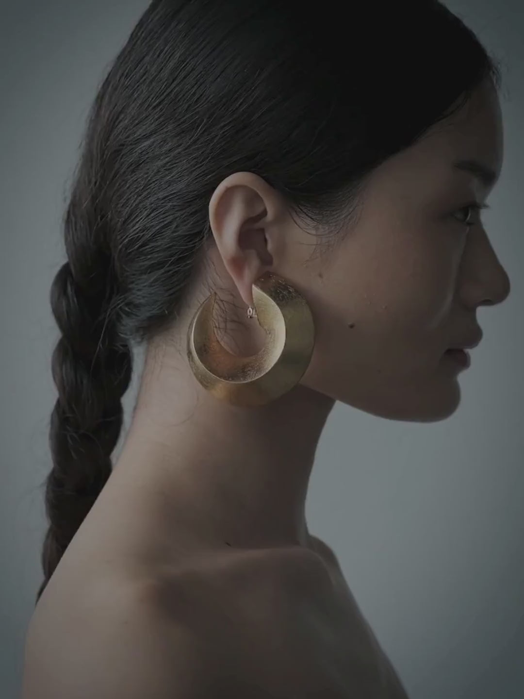 A woman in profile wearing large, chunky gold hoop earrings and a black strapless top, set against a solid slate gray background.
