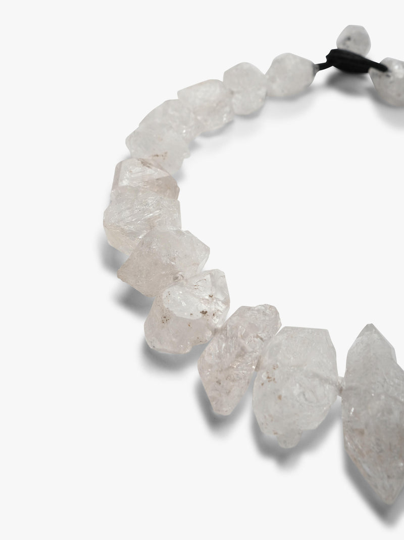 A beaded stretch necklace made of  white raw crystal chips, tied with a black elastic band, shown on a plain light gray background.