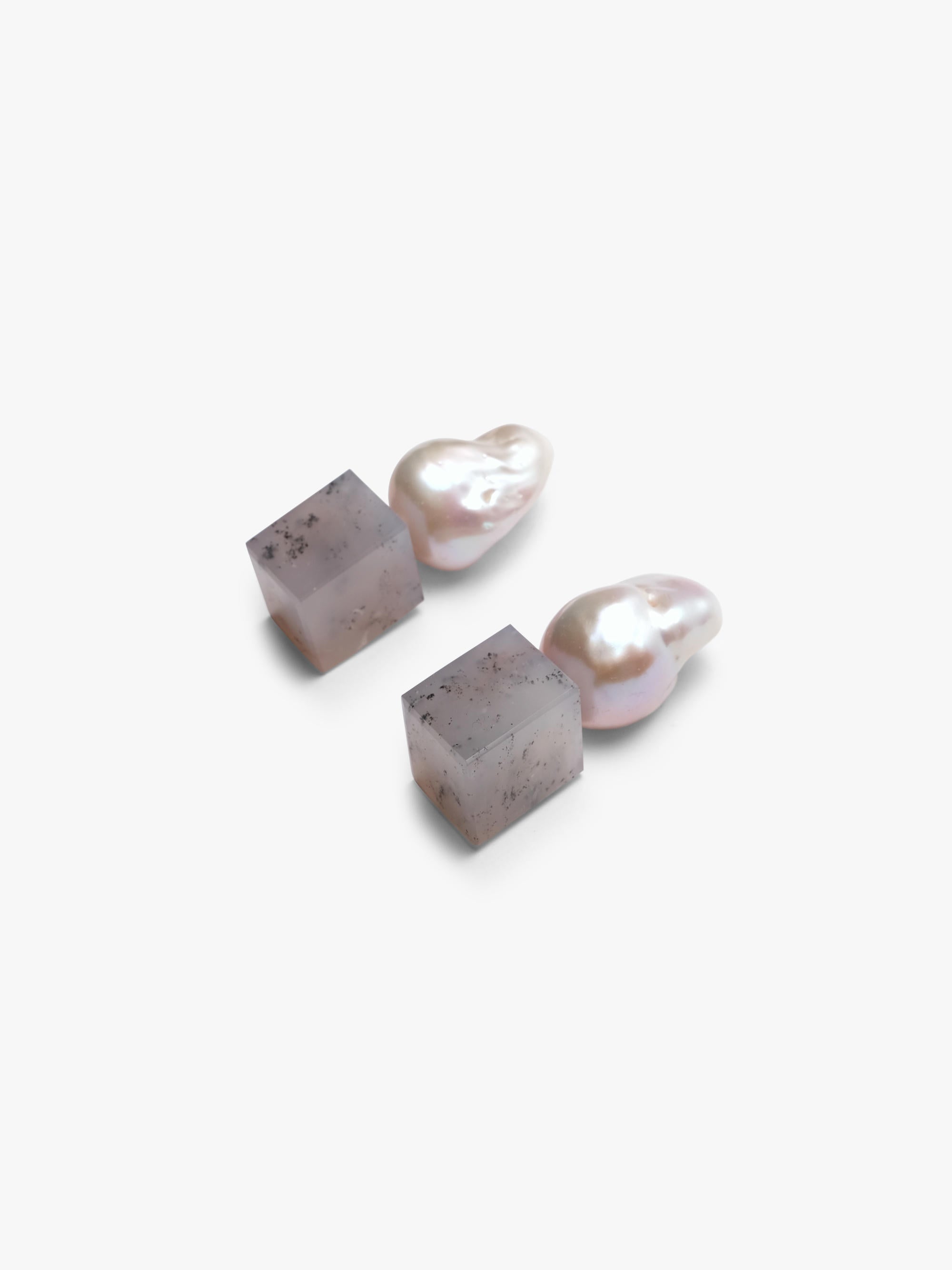 A pair of minimalist drop earrings featuring organic baroque pearls suspended from square, textured gray stone pendants, set against a clean white background.