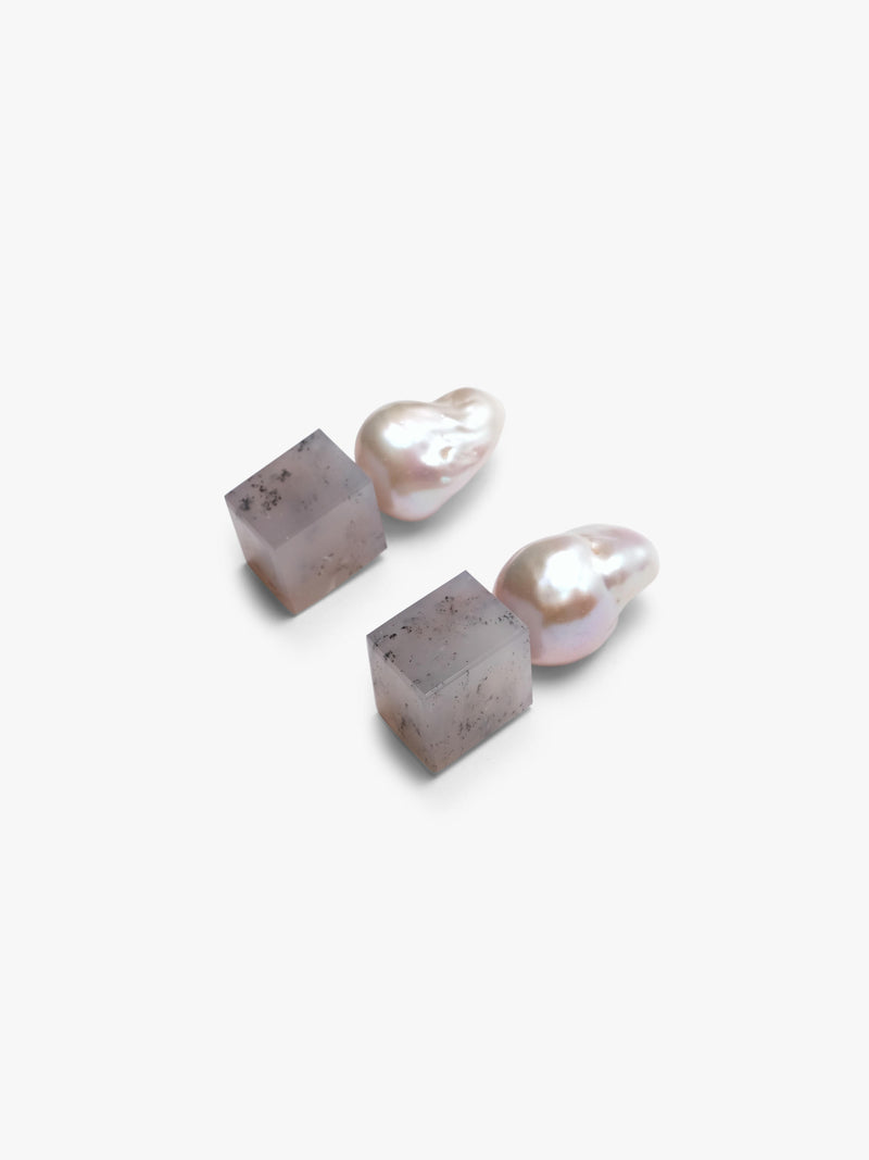 A pair of minimalist drop earrings featuring organic baroque pearls suspended from square, textured gray stone pendants, set against a clean white background.
